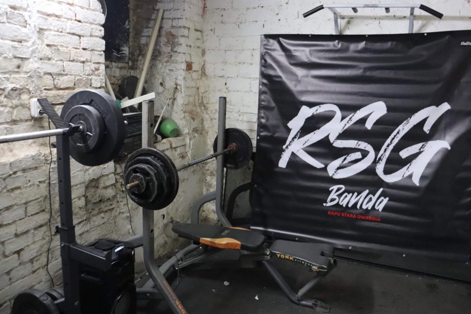 Piwnica RSG — ławka, sztangi i baner RSG Banda